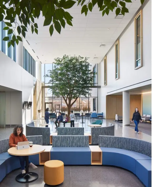 Spacious corporate atrium with indoor trees, curved lounge booths, tall clerestory windows and people gathered at a refreshment table.