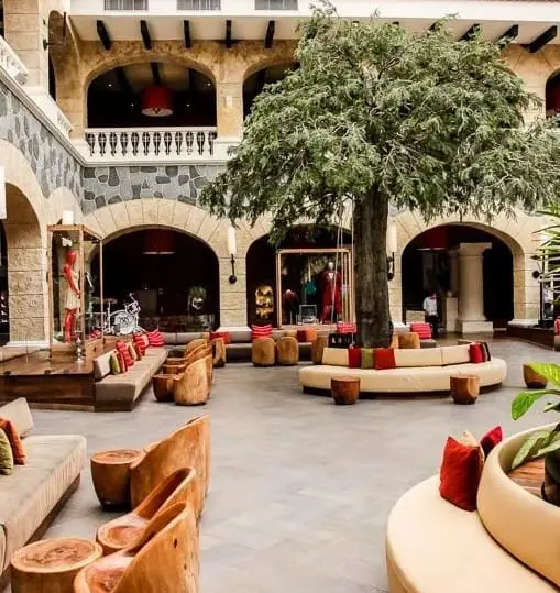 Indoor courtyard lounge with large tree, circular seating, carved wood stools and stone arches—Spanish-style boutique hotel.