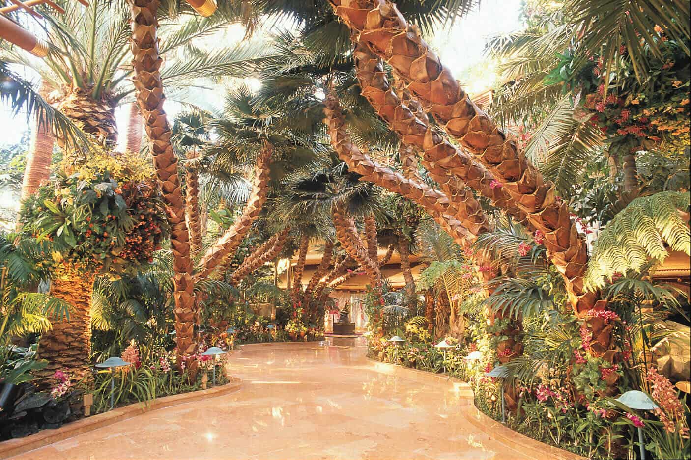 Indoor tropical atrium walkway with arching palm trees, orchids and ferns along a polished marble path—luxury hotel conservatory.