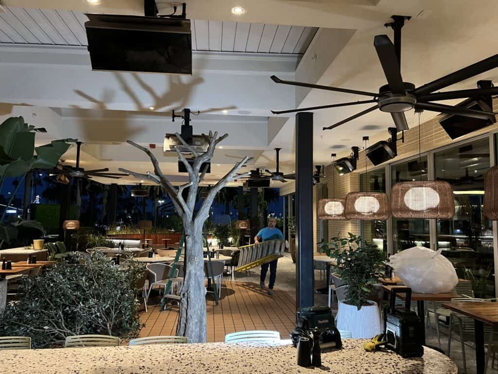 Outdoor restaurant at night with artificial tree, ceiling fans, hanging lamps, and staff carrying yellow ladder for maintenance