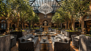 How Much Do Custom Artificial Trees Cost for Commercial Projects? Luxurious atrium courtyard featuring custom artificial olive trees and stylish gray sofas arranged around a central fountain. The elegant space is illuminated by warm ambient lighting from chandeliers and brass lanterns, complemented by lush greenery. The glass-and-metal conservatory roof overhead adds a touch of sophistication, while the intricate brick walls and carefully designed seating areas offer a cozy yet refined atmosphere. Perfect for showcasing the beauty and value of custom artificial trees in commercial interior design projects.